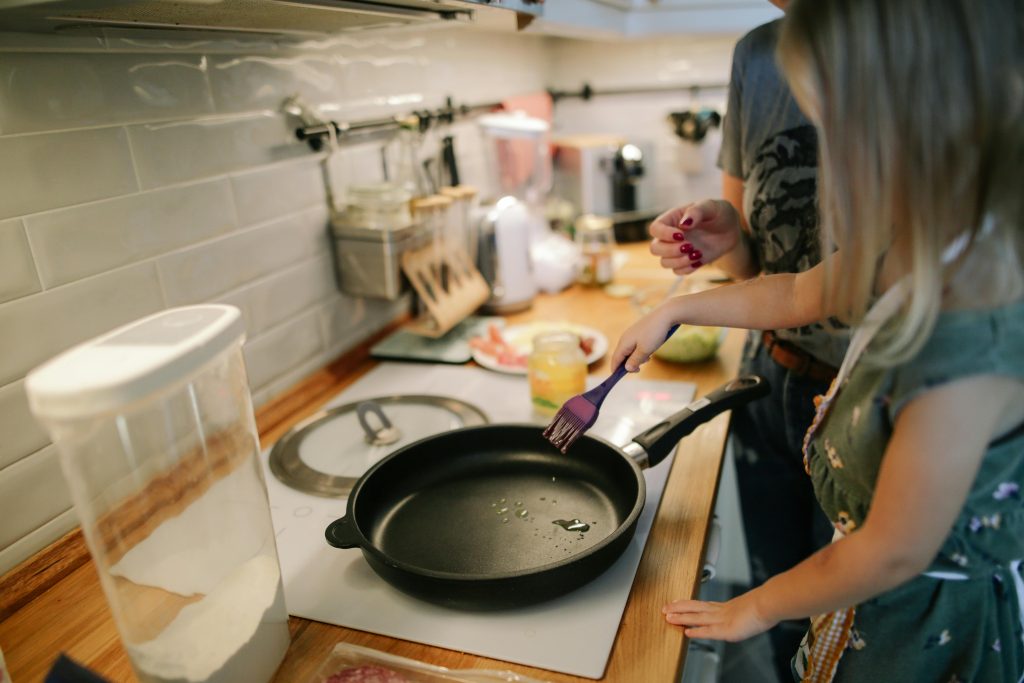 pexels-dariaobymaha-1684032 There is a kitchen bench with a stove in focus. There is a large pan on the stove and one female has spatula stirring the contents whilst another female looks on