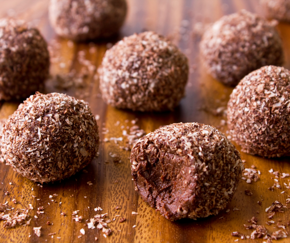 No bake bliss balls on a wooden board. They are small and round, covered in coconut and the front one has a bite taken out of it