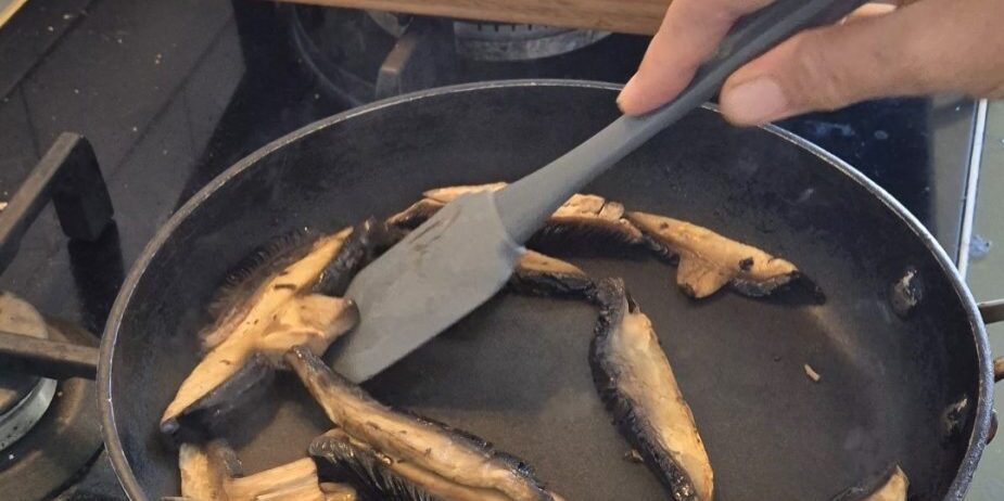 A frying pan on a stove with sliced mushrooms in it. A hand holding a spatula is moving the mushrooms around the pan. There is a wooden chopping board resting on the stove top