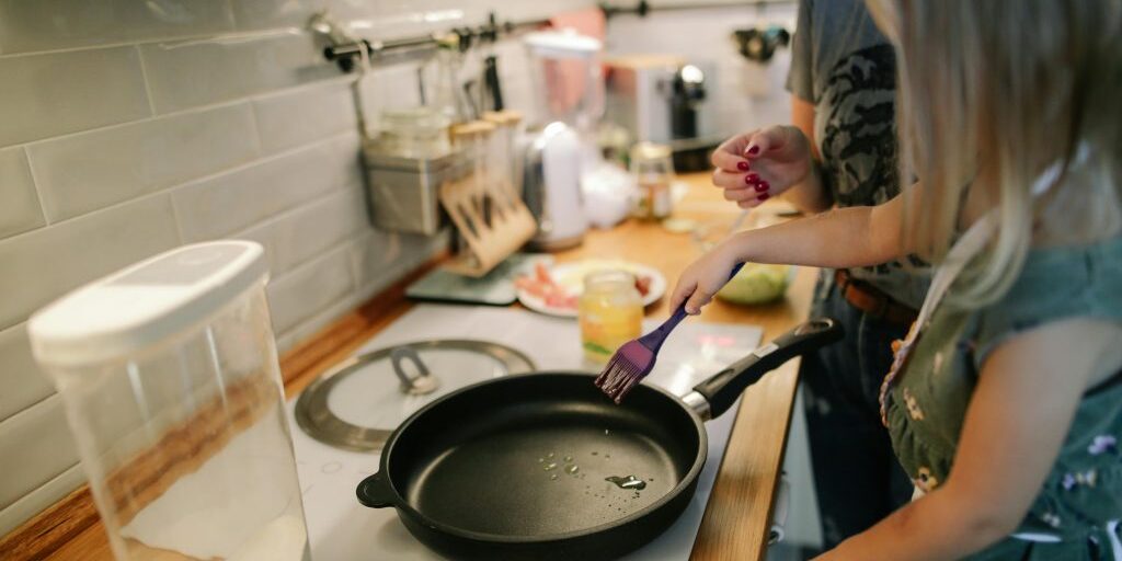 There is a kitchen bench with a stove in focus. There is a large pan on the stove and one female has spatula stirring the contents whilst another female looks on