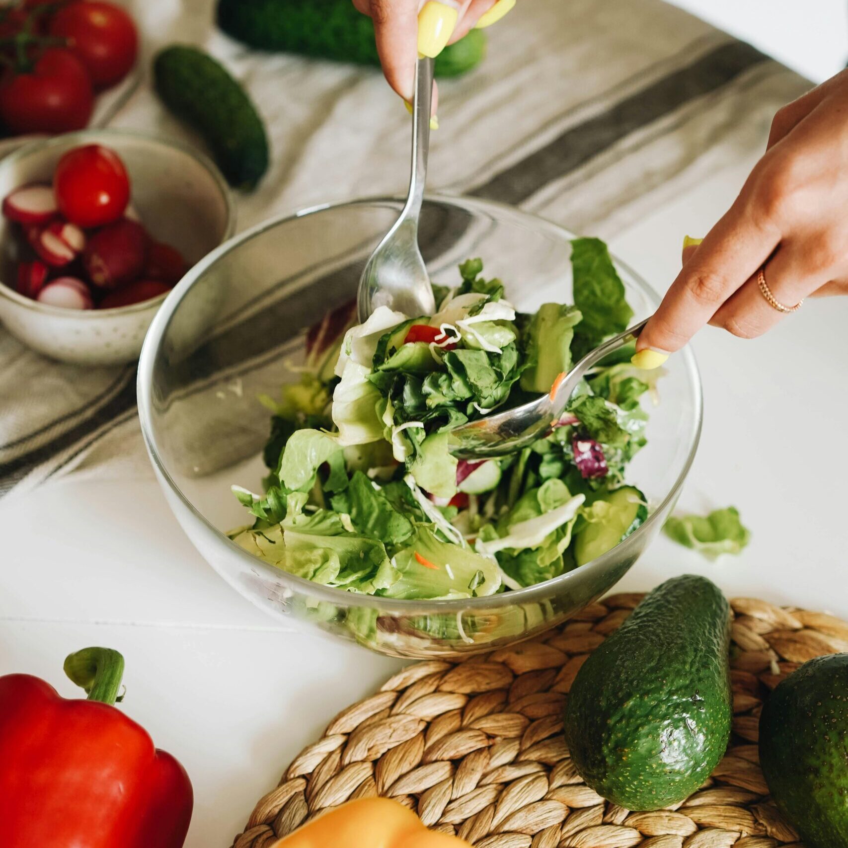 hands mixing a salad in a glass bowl with three different coloured capsicums on the bench