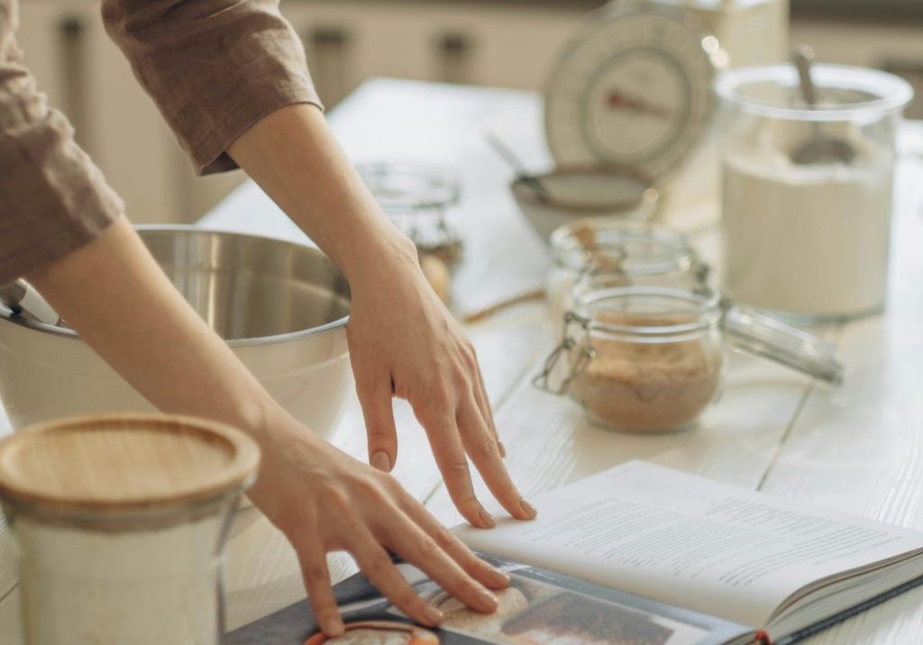 hands resting on a cookbook sitting on a bench surrounded by kitchen utensils such as bows, scales and jars