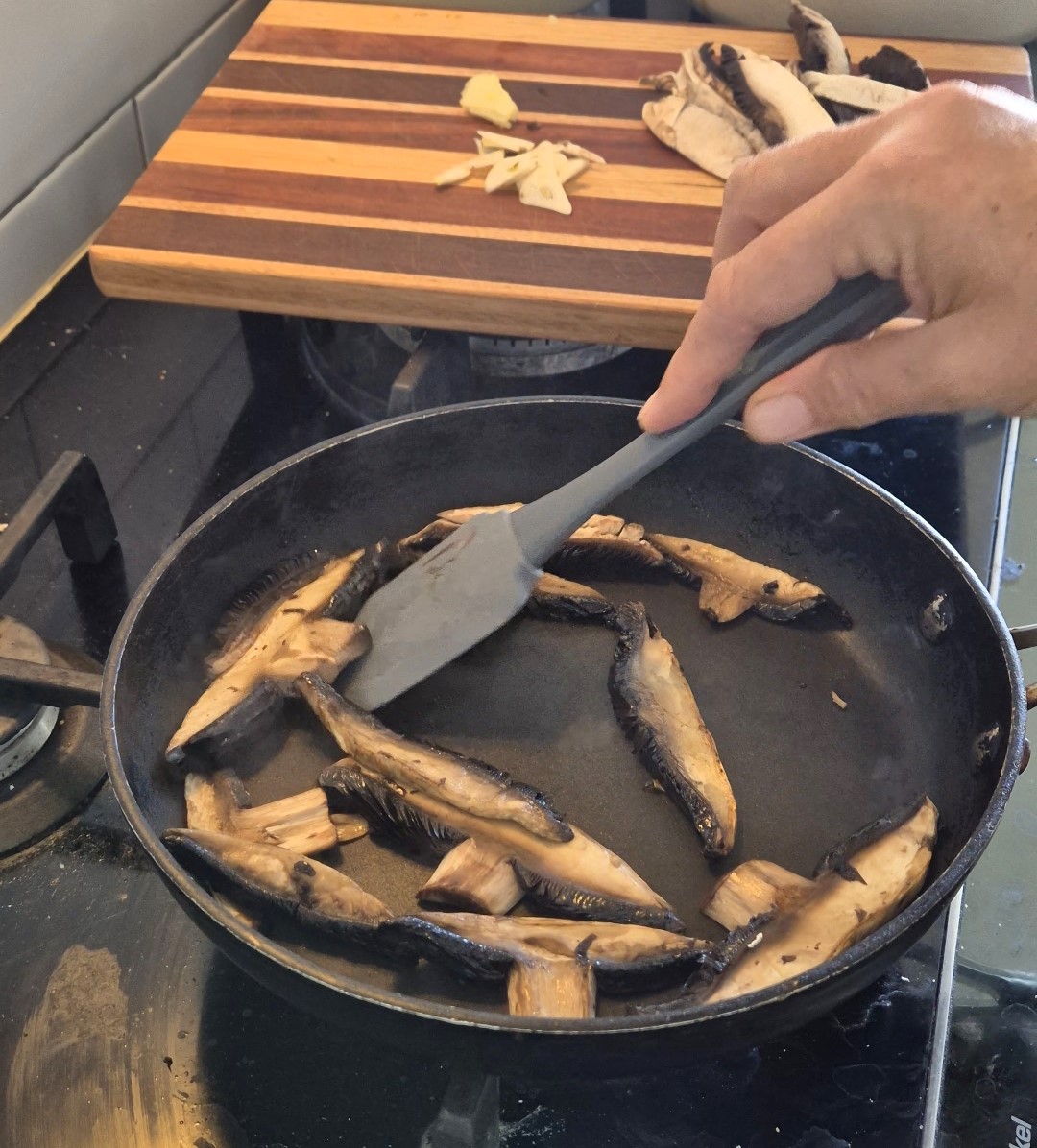 A frying pan on a stove with sliced mushrooms in it. A hand holding a spatula is moving the mushrooms around the pan. There is a wooden chopping board resting on the stove top