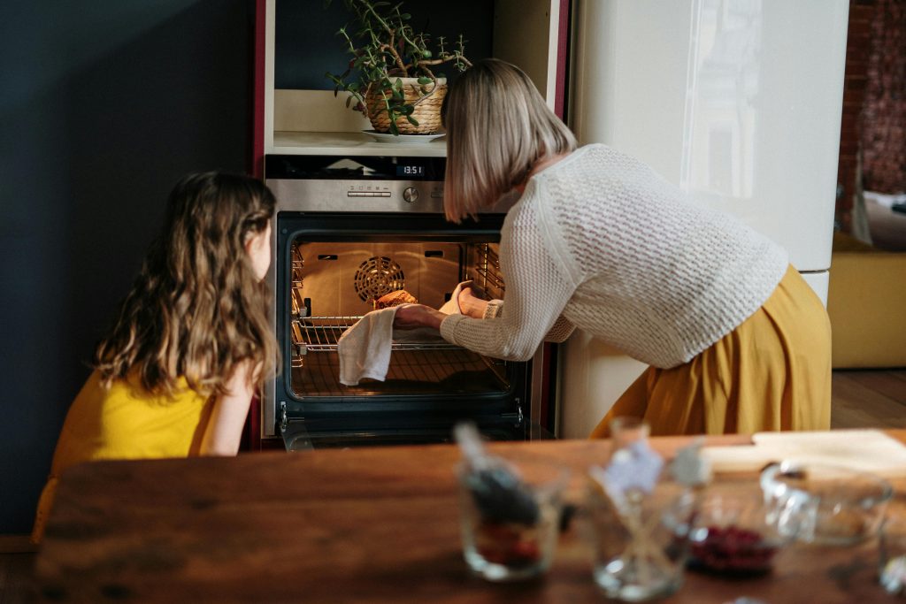 A white female and female child are standing with their backs to the camera looking at an open oven. The lady is handling a hot item in the oven with gloves on whilst the child looks on