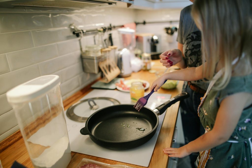 There is a kitchen bench with a stove in focus. There is a large pan on the stove and one female has spatula stirring the contents whilst another female looks on