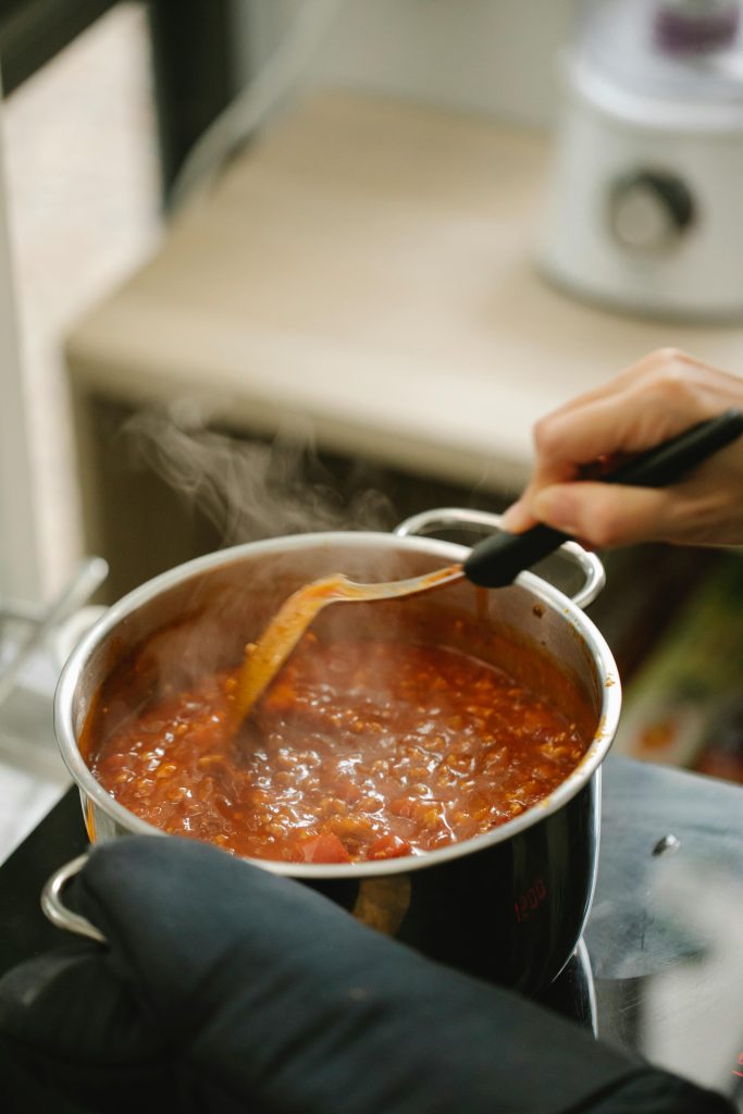 A pot of sauce that looks like bolognese or similar red casserole is on a stove top. Two hands are holding the sides, one with an oven mitt on