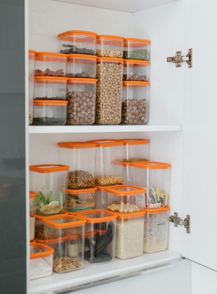 A group of containers with pantry items are sitting on white shelf. The containers are clear with wooden lids and are stacked neatly in different sizes